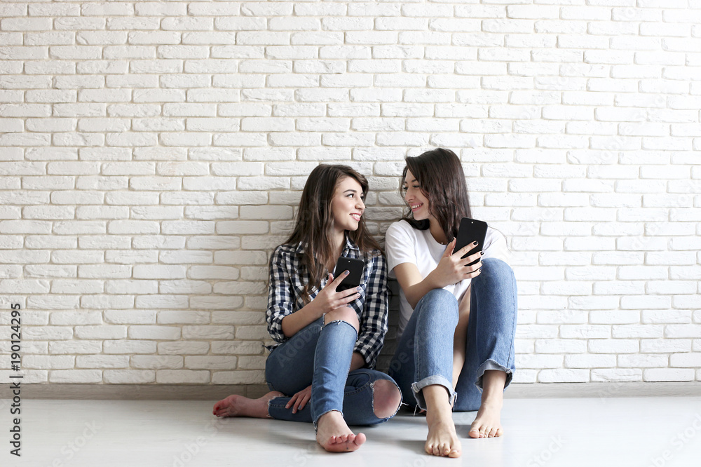 Happy millennial girls having fun indoors. Young beautiful women with perfect charismatic smiles, brown eyes, wavy dark hair. Minimalistic interior, white brick textured wall background, loft style.