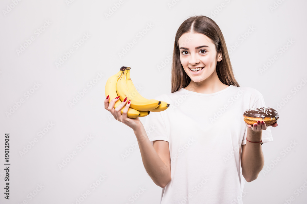 Ταπετσαρία Young healthy woman make a choise between bananas or donut isolated on white background