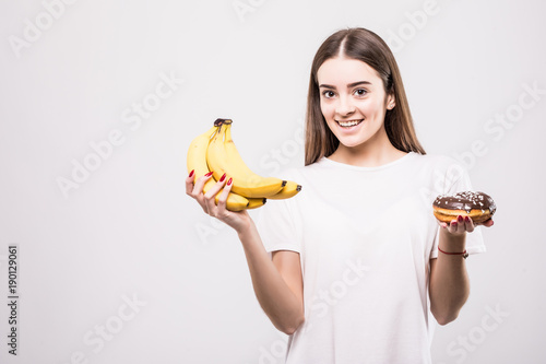 Fotografía Young healthy woman make a choise between bananas or donut isolated on white background