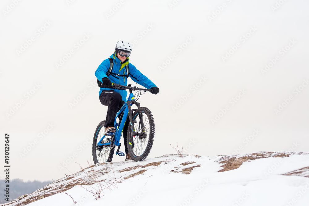 Cyclist in Blue Riding Mountain Bike on Rocky Winter Hill Covered with Snow. Extreme Sport and Enduro Biking Concept. Cyclist in Blue Riding Mountain Bike on Rocky Winter Hill Covered with Snow. Extreme Sport and Enduro Biking Concept.
