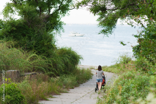 Young woman walking at sea background with little cute child baby boy in baby carriage on nature, green trees. Mother, little kid son. Parenthood, family day 15 of may, love, parents, children concept