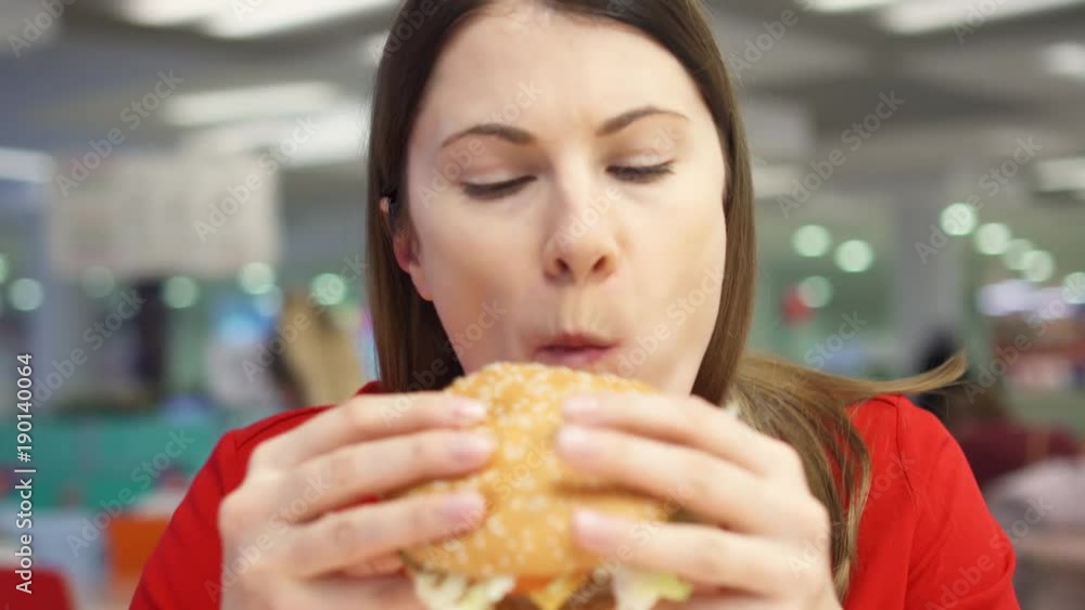 Hungry girl eating hamburger on food court. Woman biting cheeseburger at fast food restaurant Hungry girl eating hamburger on food court. Woman biting cheeseburger at fast food restaurant