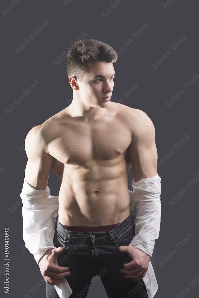 Handsome young muscular man shirtless wearing jeans, taking off leather jacket on naked muscle torso, on dark background in studio shot