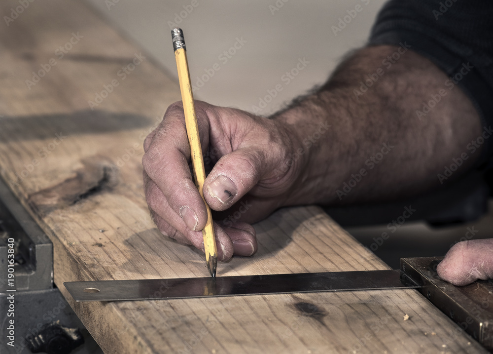 Closeup of carpenter's rough hands using a pencil and old square to mark a line on a wood board to cut using a chop saw