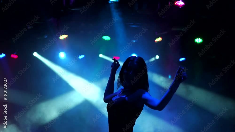 Silhouette of a slender dancing girl in a nightclub. Sexy girl dances in the dark with the light of multi-colored searchlights, slow movement. Silhouette of a slender dancing girl in a nightclub. Sexy girl dances in the dark with the light of multi-colored searchlights, slow movement.