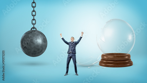 A small victorious businessman stands near a broken glass sphere and an iron wrecking ball.