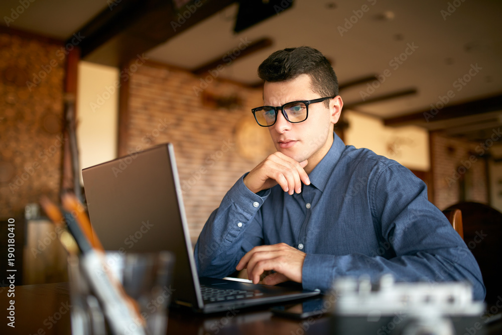 Thoughtful young caucasian businessman in glasses working on laptop computer. Pensive attractive hipster freelancer thinks over the solution of the problem with computer in cafe. Business concept. Thoughtful young caucasian businessman in glasses working on laptop computer. Pensive attractive hipster freelancer thinks over the solution of the problem with computer in cafe. Business concept.