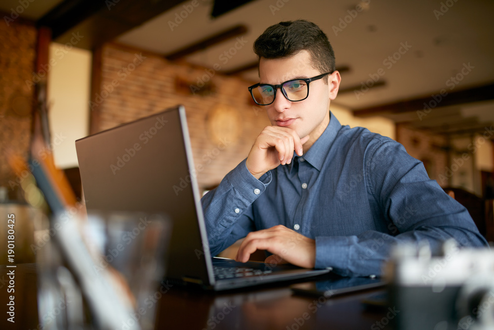 Thoughtful young caucasian businessman in glasses working on laptop computer. Pensive attractive hipster freelancer thinks over the solution of the problem with computer in cafe. Business concept. Thoughtful young caucasian businessman in glasses working on laptop computer. Pensive attractive hipster freelancer thinks over the solution of the problem with computer in cafe. Business concept.