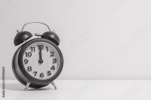 Closeup alarm clock for decorate in 12 o'clock on white wood desk and cream wallpaper textured background in black and white tone with copy space