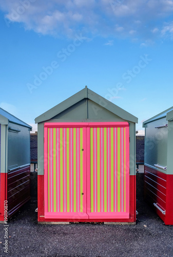 a beach hut with a multi coloured door, stripey yellow, pink, colours, in the foreground is asphalt in the background blue sky