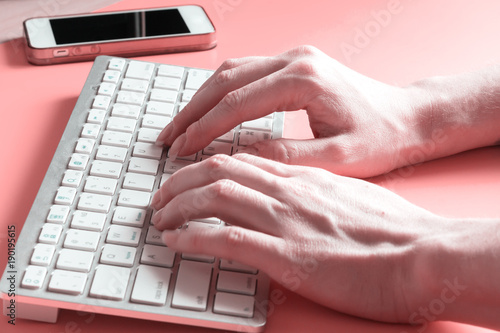 The girl's hands with the white keyboard and smartphone. Technology close-up background with laptop and pink table.