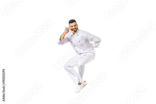 young male chef tasting food with spoon while jumping and smiling at camera isolated on white