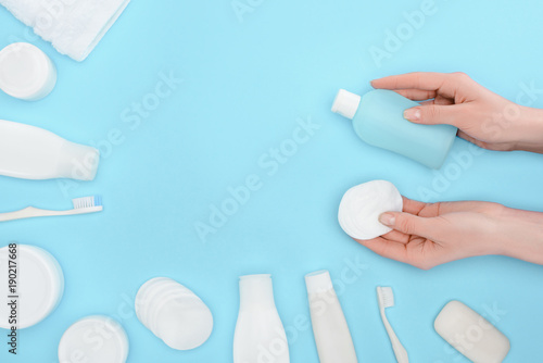 cropped view of woman holding cotton swab and lotion isolated on blue with toothbrushes, soap and cream