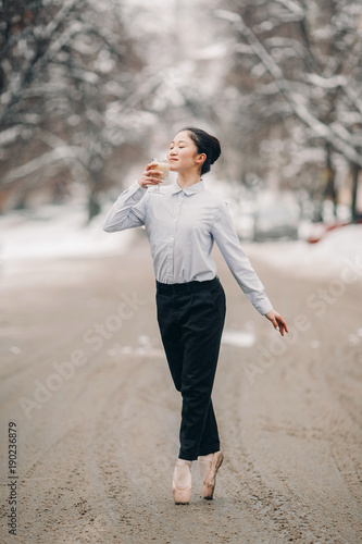 Ballerina with glass of hot coffee raf in her hands is standing at city street against snow and winter background. © Stanislav