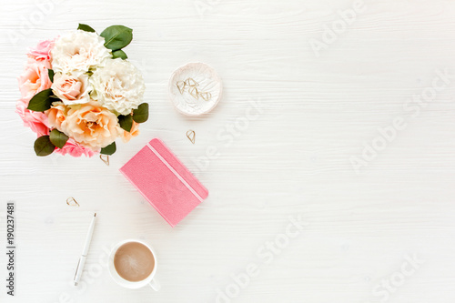 Flat lay home office desk. Female workspace with pink and beige roses flowers bouquet, coffee and chocolates on white wooden background. Top view feminine background.