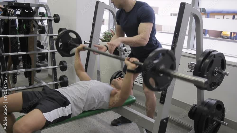 Young athlete is doing bench press with help of trainer in gym. Man raises free weight under control of instructor in sports club. Handsome guy lies on bench and does exercise with iron object Young athlete is doing bench press with help of trainer in gym. Man raises free weight under control of instructor in sports club. Handsome guy lies on bench and does exercise with iron object