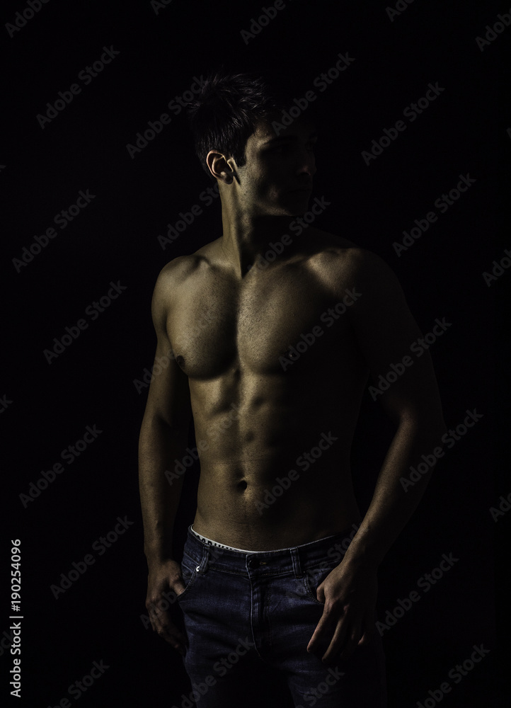 Handsome, fit muscular young man shirtless, wearing only jeans standing on black background, looking away to a side