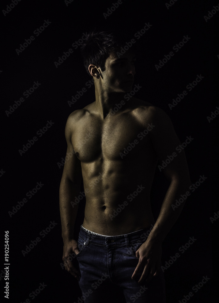 Handsome, fit muscular young man shirtless, wearing only jeans standing on black background, looking away to a side Handsome, fit muscular young man shirtless, wearing only jeans standing on black background, looking away to a side
