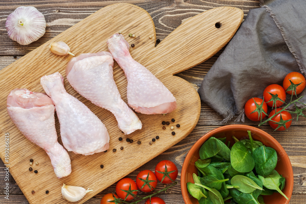 Raw chicken drumstick on a cutting board, cherry tomatoes, spinach, garlic, towel on a wooden table, top view. Raw chicken drumstick on a cutting board, cherry tomatoes, spinach, garlic, towel on a wooden table, top view.