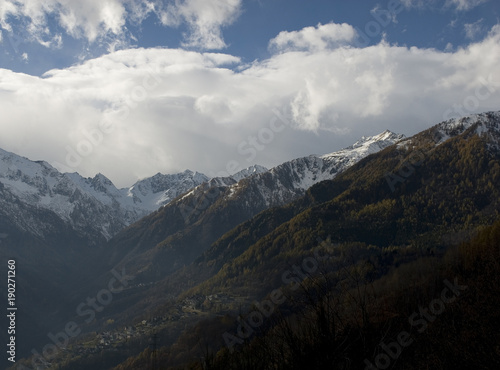 landscape: silhouette of mountains, Antrona Valley, near Swiss, snow on peaks, colored larch forest, sunset, ray of light, Alps, blue sky, clouds, village, late autumn, winter, Piedmont, Italy