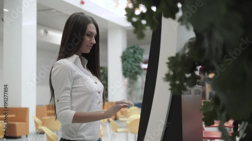 Caucasian female using automated teller machine with big digital screen while standing in shopping mall, woman verifies account balance on banking application via modern device icon photo
