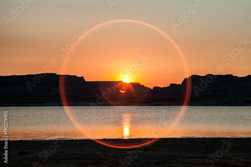 Sunrise with sun flare circle on lake with silhouetted plateaus.