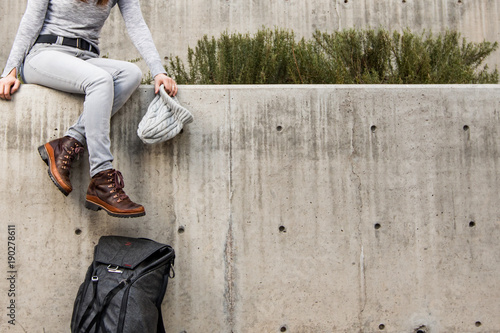 Woman dangling legs off the edge of a concrete ledge with grey beanie hat in hand and  backpack leaning against wall. Lots of negative space to the right.