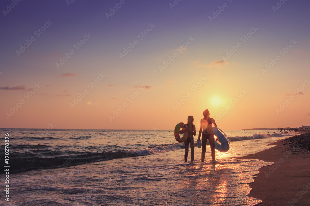 Two young girls walk along sea beach at sunset behind them. Summer vacation at the sea. A company of teenagers is bathing in sea with inflatable circles. Summer vacation at sea concept.