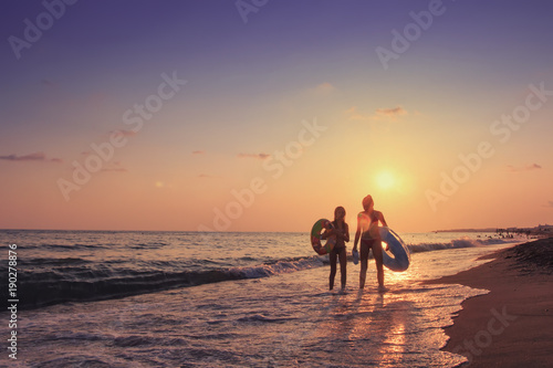Two young girls walk along sea beach at sunset behind them. Summer vacation at the sea. A company of teenagers is bathing in sea with inflatable circles. Summer vacation at sea concept.