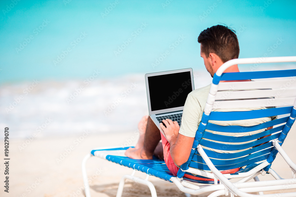 Young man with laptop on tropical caribbean beach. Man sitting on the sunbed with computer and working on the beach