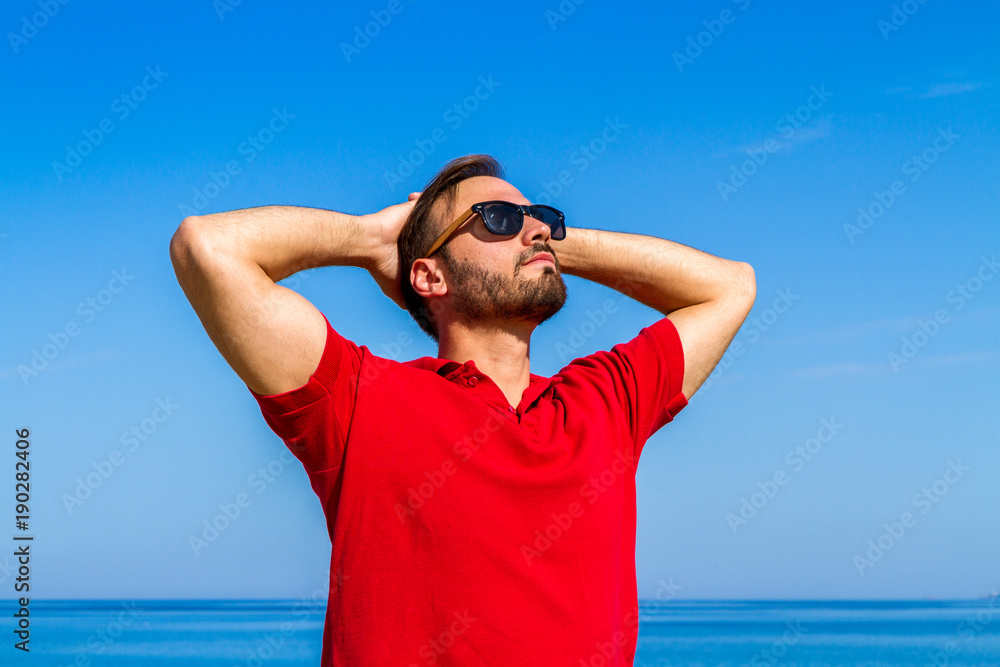 Dynamic, ambitious, successful, expressive bearded young in red t-shirt sunglasses in summer blue sky ocean background. Self-confidence lifestyle concept Dynamic, ambitious, successful, expressive bearded young in red t-shirt sunglasses in summer blue sky ocean background. Self-confidence lifestyle concept