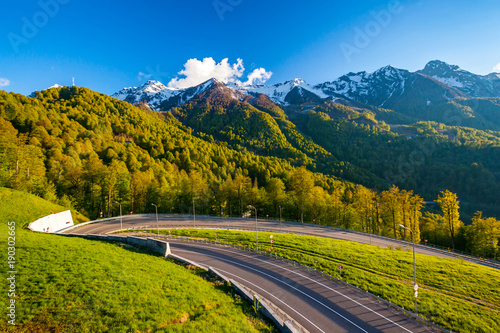 A winding modern asphalt road to the top of a resort in the bright yellow rays of the evening sun, dense forest and the Caucasian mountain range. Summer spring forest mountain landscape, Sochi Russia.