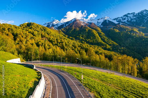 A winding modern asphalt road to the top of a resort in the bright yellow rays of the evening sun, dense forest and the Caucasian mountain range. Summer spring forest mountain landscape, Sochi Russia.