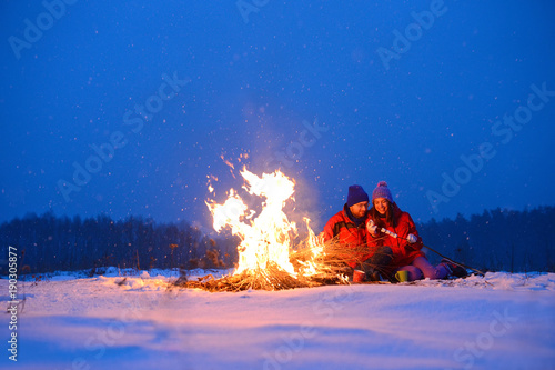  Happy loving couple sitting at a fire in the snow among the fields in the winter against the background of the forest and the sky on Valentine's Day.