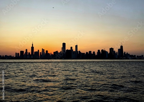 Evening sunset over Chicago silhouettes the skyline, as seen from Lake Michigan, which is glowing with orange sunlight.