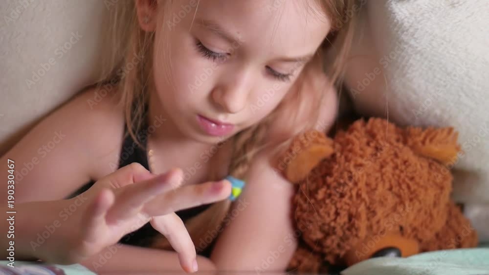 Cute little girl sitting on bed together with teddy bear playing games on tablet. Cute little girl sitting on bed together with teddy bear playing games on tablet.
