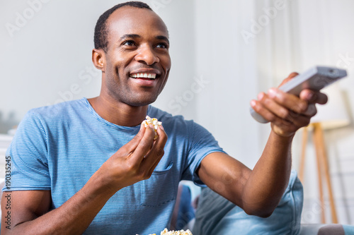 Leisure time. Happy joyful adult man holding a remote control and eating popcorn while choosing a TV channel