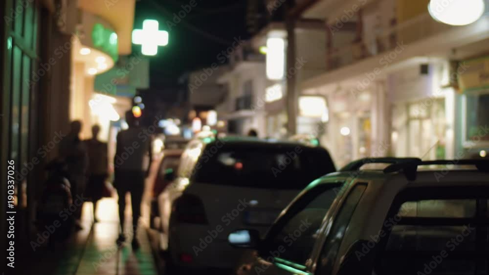 Street in the town at night illuminated with outside banners. Cars parked alongside, people walking on sidewalk Street in the town at night illuminated with outside banners. Cars parked alongside, people walking on sidewalk