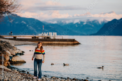 One happy kid girl playing by lake Geneva at sunset with swiss mountains Alps on background. Image taken in Lausanne, Switzerland