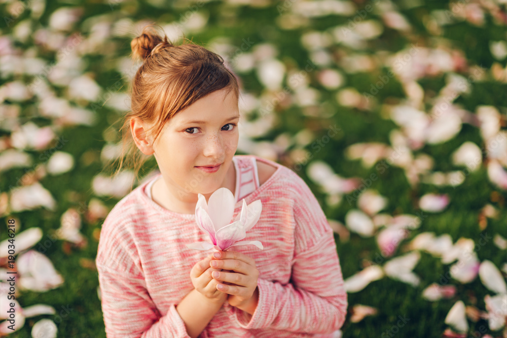 Spring portrait of cute little girl sitting on green grass with flowers petals, holding beautiful Magnolia flower, wearing pink sweatshirt, fashion for children