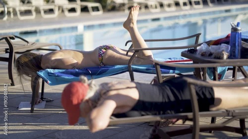 Muscular pretty woman in bikini, sunglasses and red hat reads a book while in a chaise lounge chair. photo