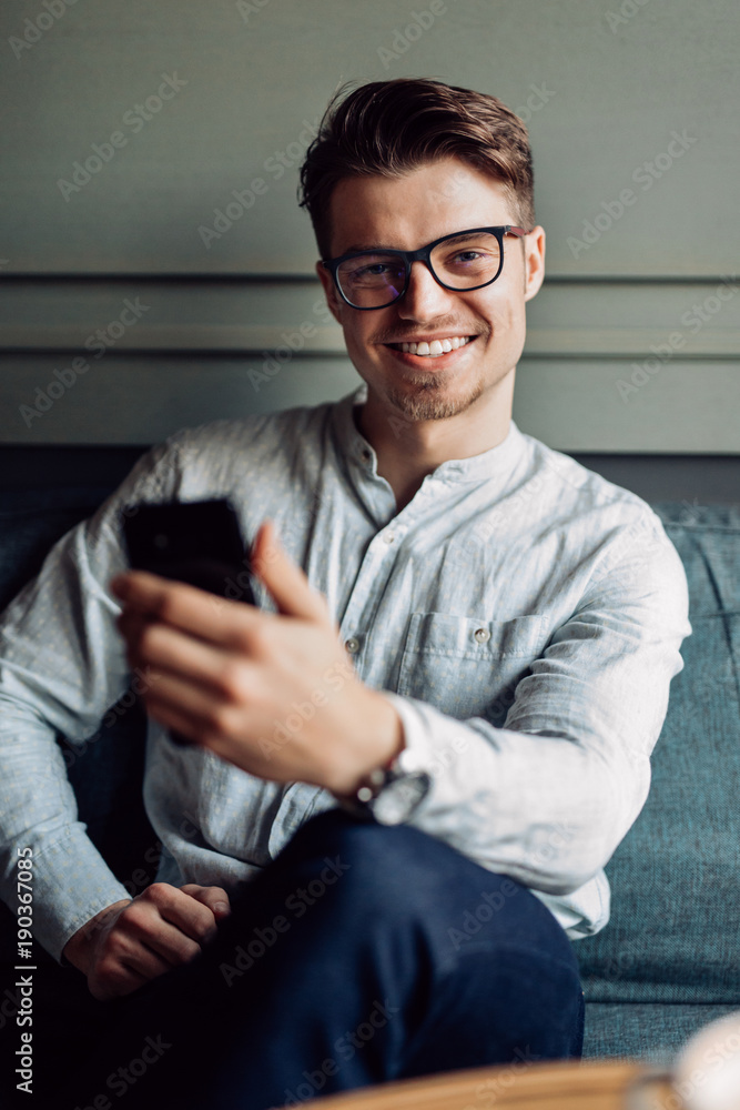 Cheerful young man in eyeglasses, holding mobile phone, sitting on couch and looking at camera. Dressed in stylish shirt and pants. Cheerful young man in eyeglasses, holding mobile phone, sitting on couch and looking at camera. Dressed in stylish shirt and pants.