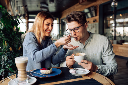Beautiful smiling girl feeds her handsome boyfriend  eating tasty cake and drinking coffee  spending time together at cafe.