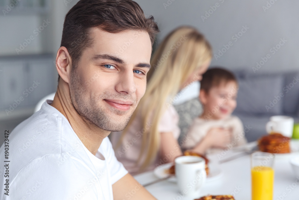 handsome young man looking at camera while her wife and kid having breakfast handsome young man looking at camera while her wife and kid having breakfast