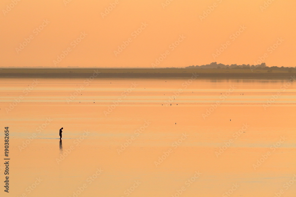 Silhouettes of man in shallow water in lake at orange red sunset