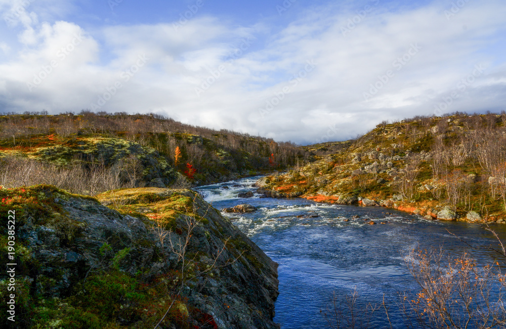 Beautiful autumn sunny landscape with a river between rocks. Bright autumn colors and trees without leaves against a blue sky with clouds Beautiful autumn sunny landscape with a river between rocks. Bright autumn colors and trees without leaves against a blue sky with clouds