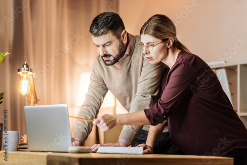 Look here. Attentive male person leaning on table and looking downwards while standing near his colleague