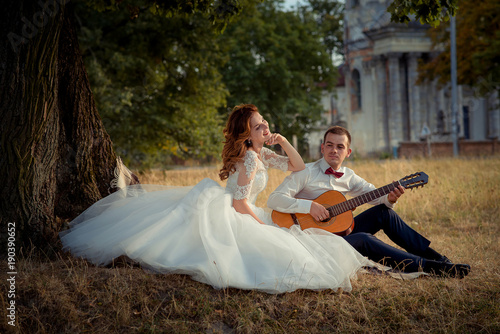 Charming smiling bride is listening to the elegant groom playing the guitar while sitting on the grass under the tree. photo