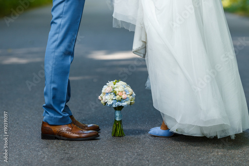 Wedding details: classic brown and blue shoes of bride and groom. Bouquet of roses standing on the ground between them. Newlyweds standing in front of each other