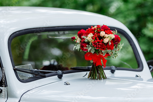 Passion wedding bouquet with dark red and marsala roses, greenery standing on white car. Bridal flowers, decorated with ribbon.